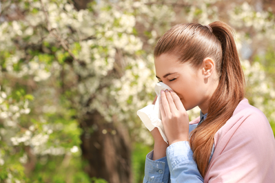 woman with seasonal allergies sneezing into tissue next to blooming tree