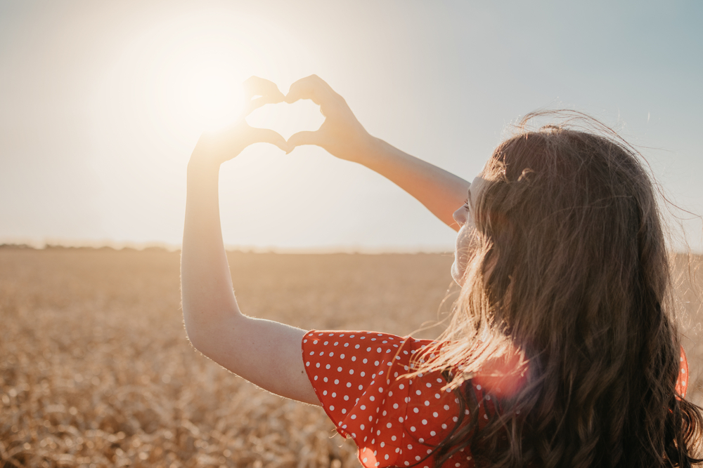 young woman making hand heart against the sun